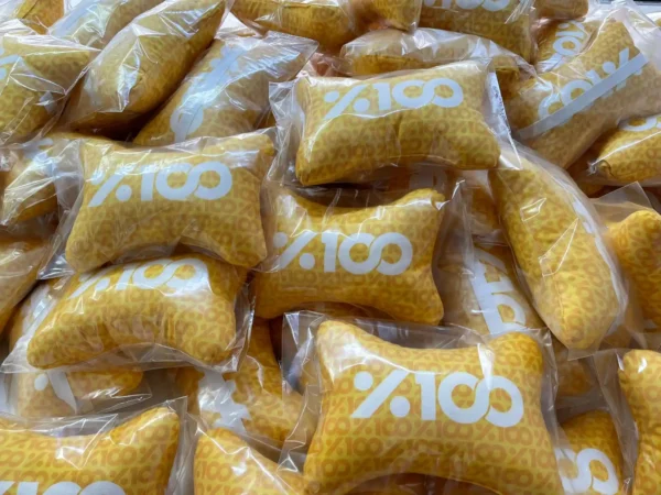 Bulk display of yellow bone-shaped neck pillows with %100 logo in transparent packaging – Textile Promos