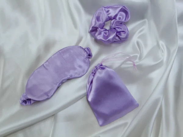 Lavender satin sleep set with eye mask, scrunchie, and pouch on white satin fabric background